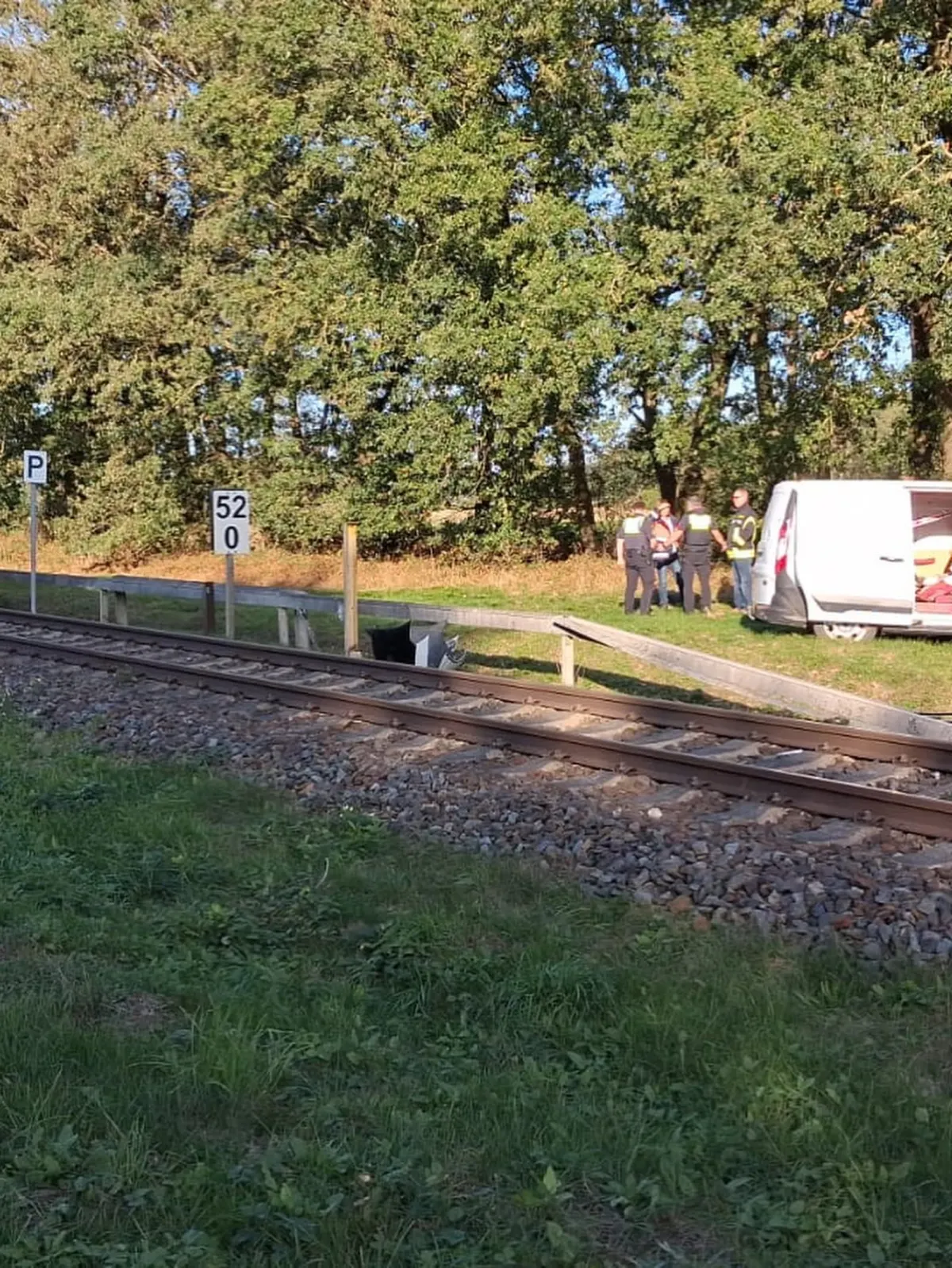Eine Gruppe von Personen in Sicherheitswesten steht neben einem weißen Van an Bahngleisen in einer ländlichen Umgebung mit Bäumen im Hintergrund.