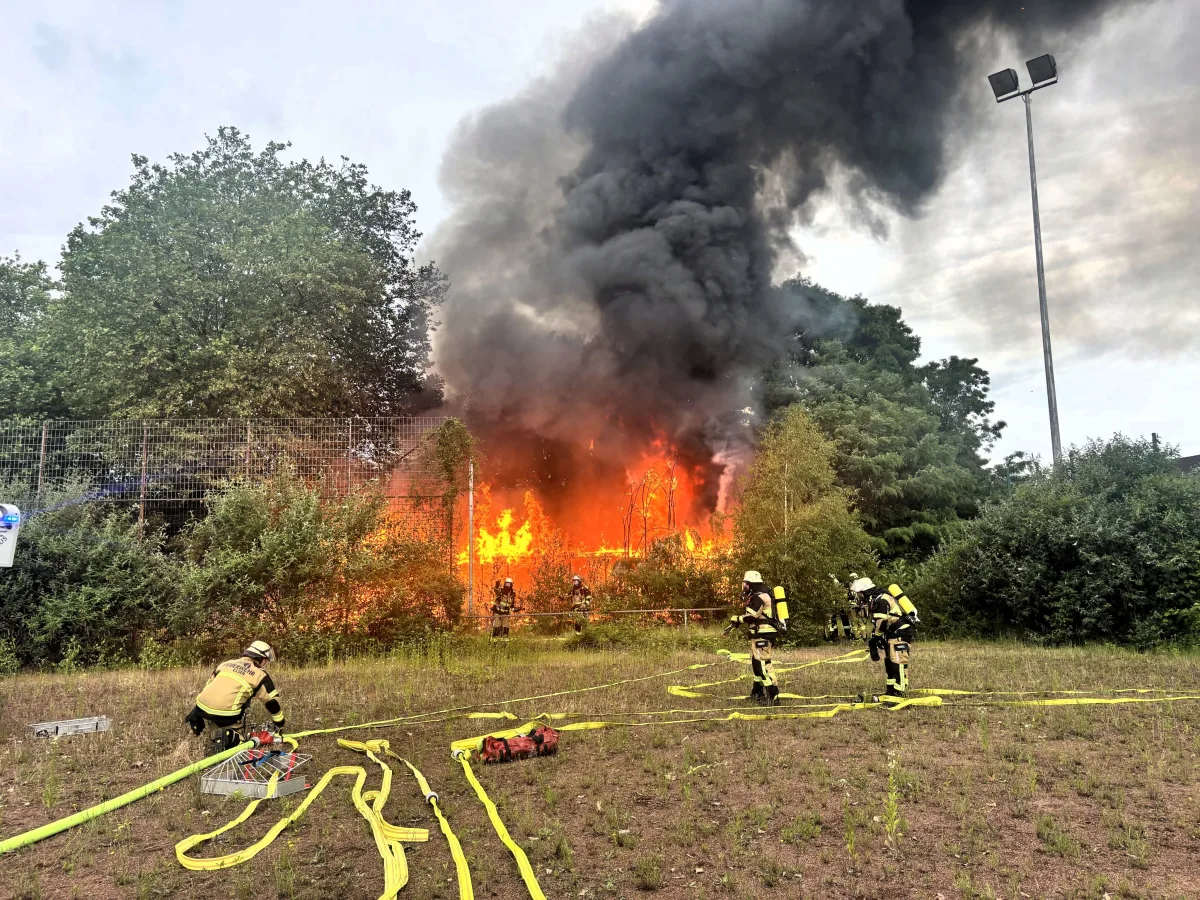 Feuerwehrleute in Schutzkleidung bekämpfen einen großen Brand im Freien. Dicker schwarzer Rauch steigt in den Himmel auf.