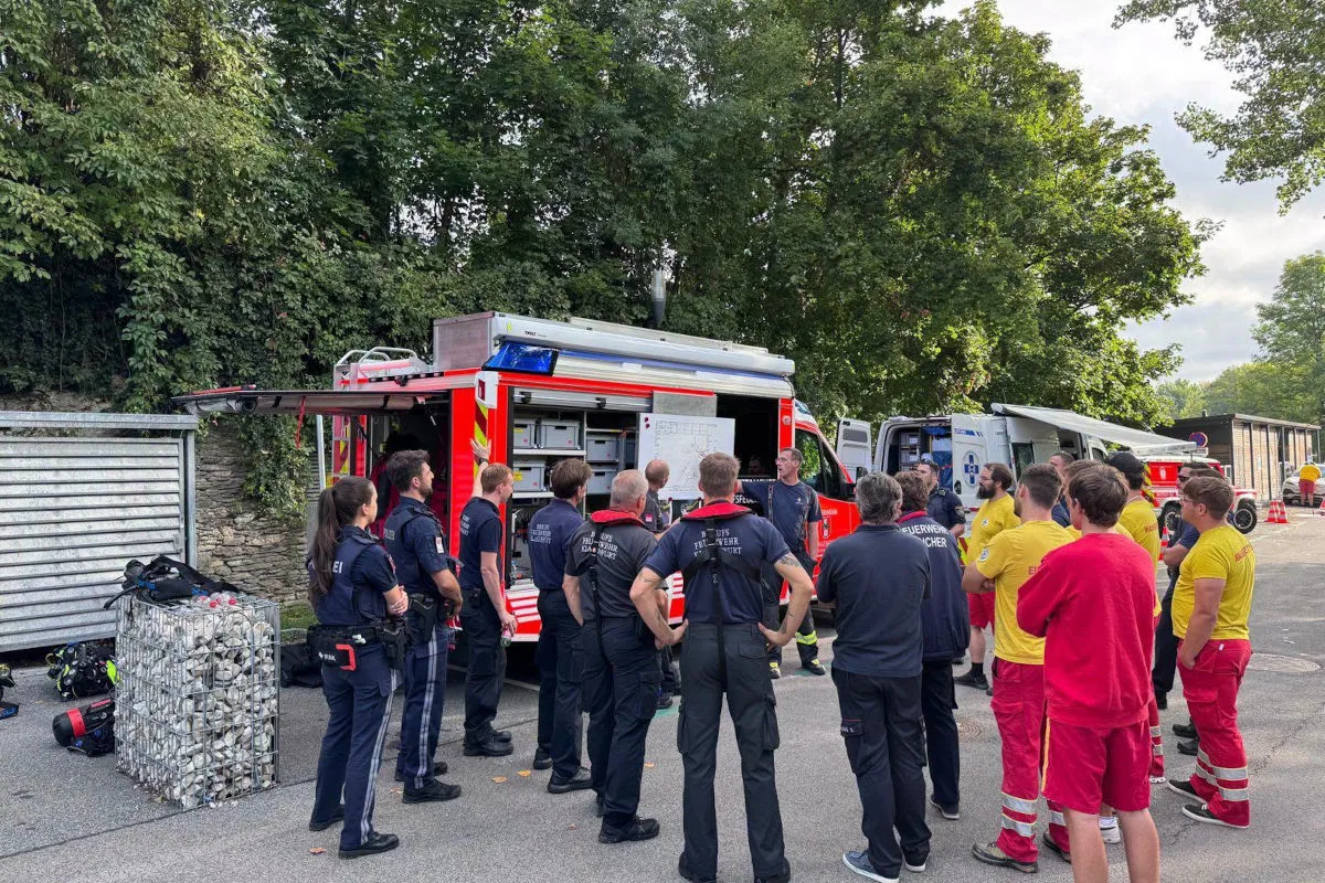 Mehrere Feuerwehr- und Rettungskräfte stehen vor einem roten Einsatzfahrzeug und beraten sich; umgeben von Bäumen.