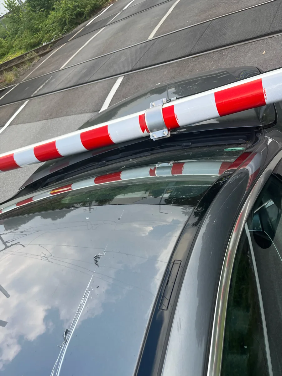 Das Bild zeigt ein graues Auto an einem Bahnübergang mit einer roten und weißen Bahnschranke, die auf dem Autodach liegt. Der Himmel ist teilweise bewölkt.
