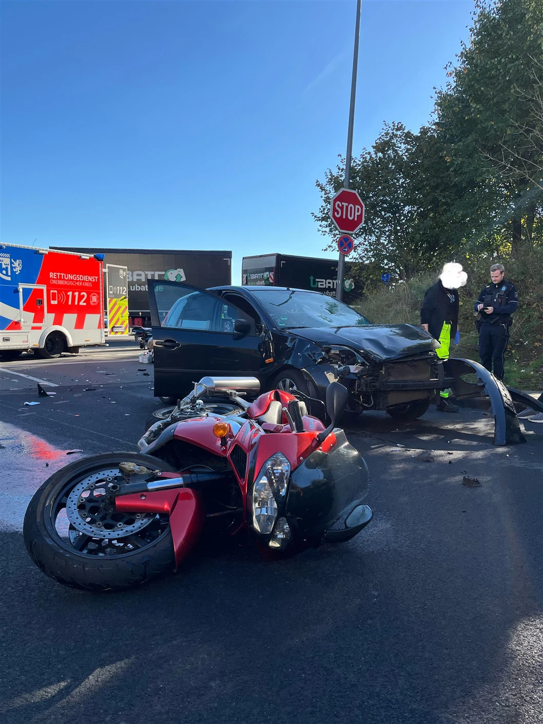 Ein rotes Motorrad liegt auf der Straße, während im Hintergrund ein stark beschädigtes schwarzes Auto und ein Krankenwagen sichtbar sind.