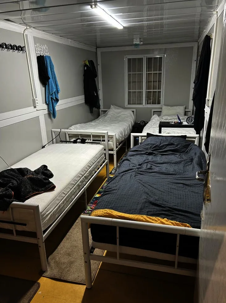 Der Schlafsaal zeigt drei Metallbetten. Auf einem Tisch liegen persönliche Gegenstände. Kleidung hängt an der Wand.