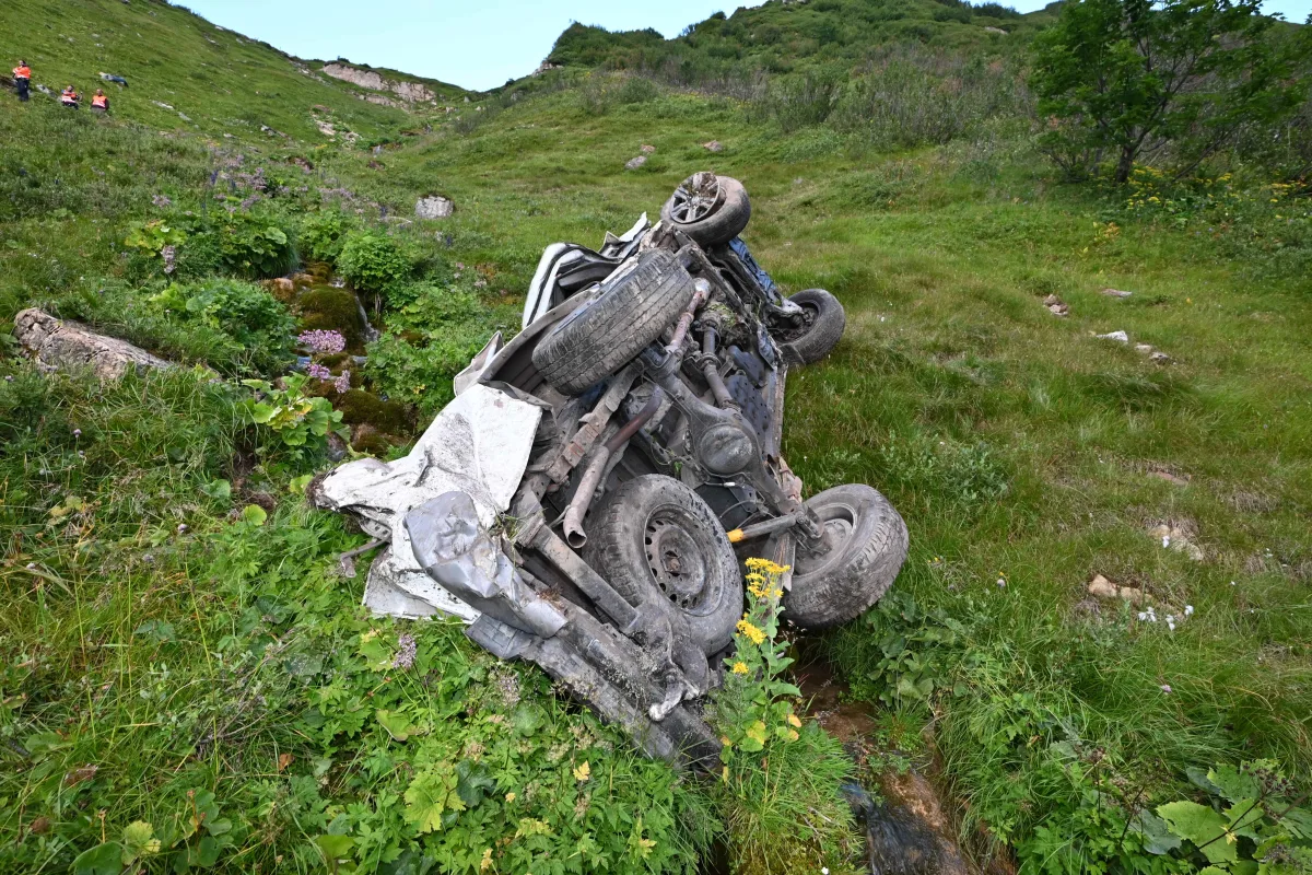 Ein verunglücktes Fahrzeug liegt seitlich gekippt in einer grasbewachsenen Umgebung an einem Hang, mit zum Teil zerstörten Karosserieteilen.