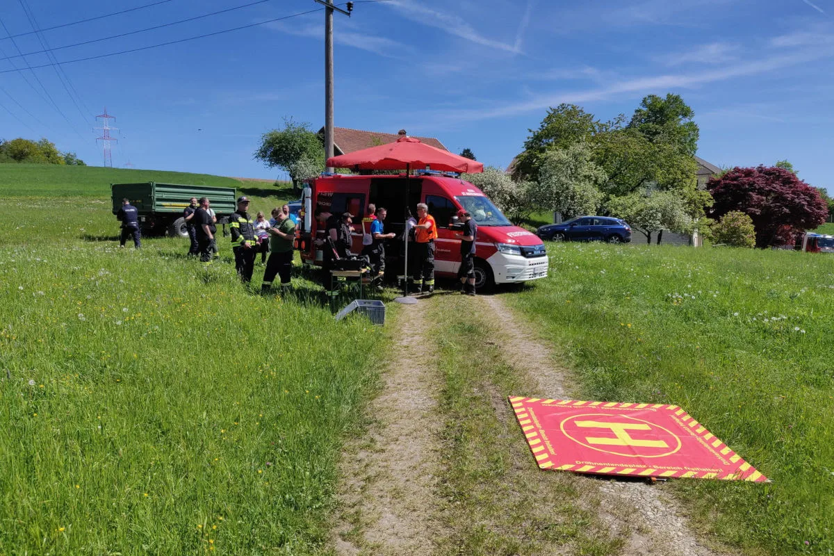 Ein rotes Einsatzfahrzeug steht auf einem Feldweg, umgeben von Menschen in Einsatzkleidung. Im Hintergrund ist grüne Landschaft und blauer Himmel zu sehen.