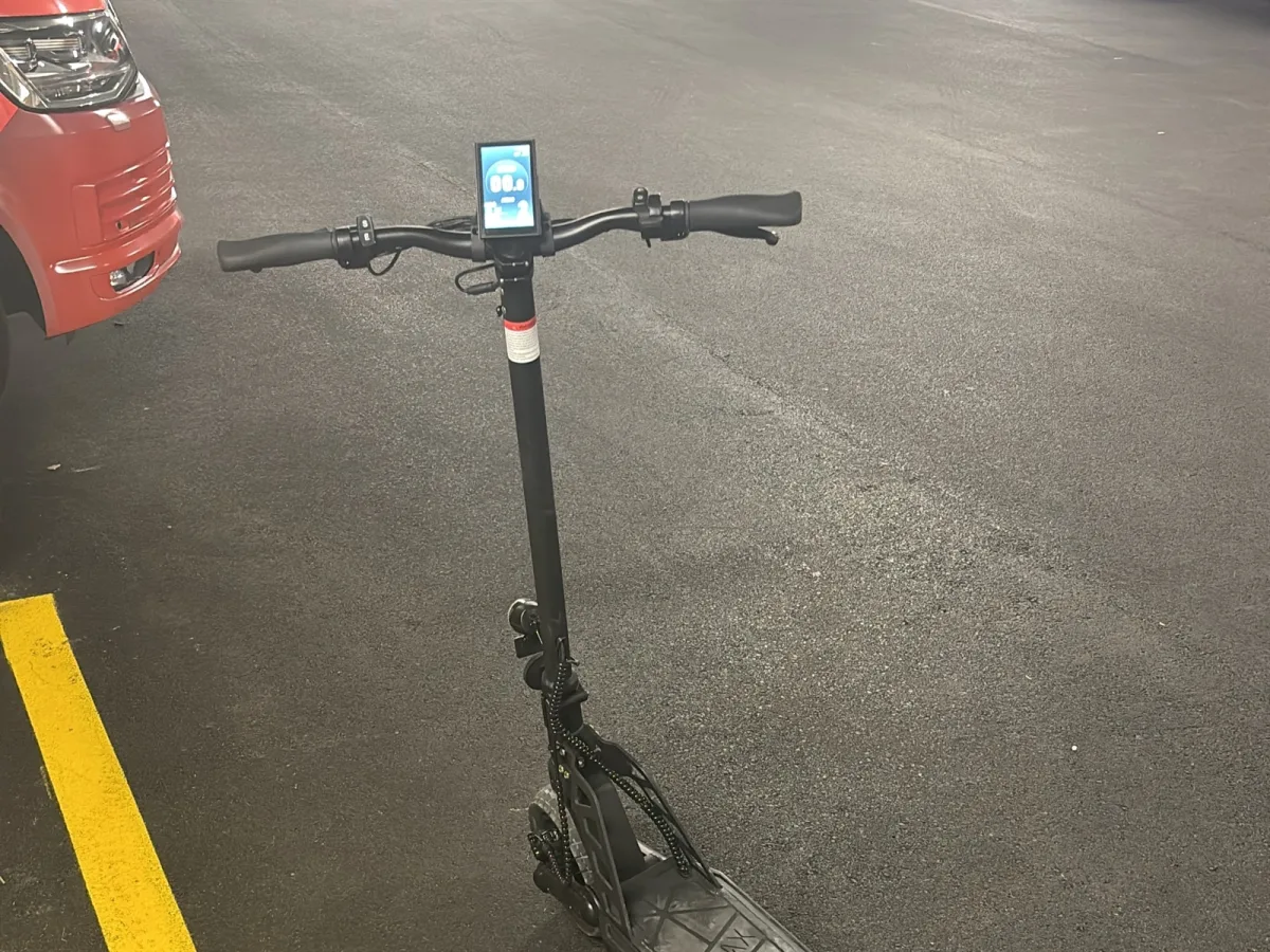 Ein E-Roller steht allein in einer Parkgarage neben einem roten Fahrzeug auf schwarzem Asphaltboden. Der E-Roller ist nach vorne gerichtet.