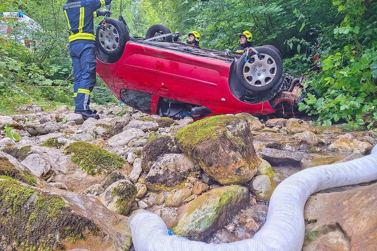 Das Bild zeigt ein rotes Auto, das auf dem Dach in einem steinigen Bachbett liegt, während Feuerwehrleute es sichern.