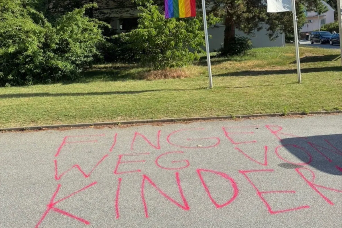 Auf dem Gehweg steht in rot 'FINGER WEG VON KINDERN', darüber hängen eine Regenbogenflagge und eine weiße Flagge.