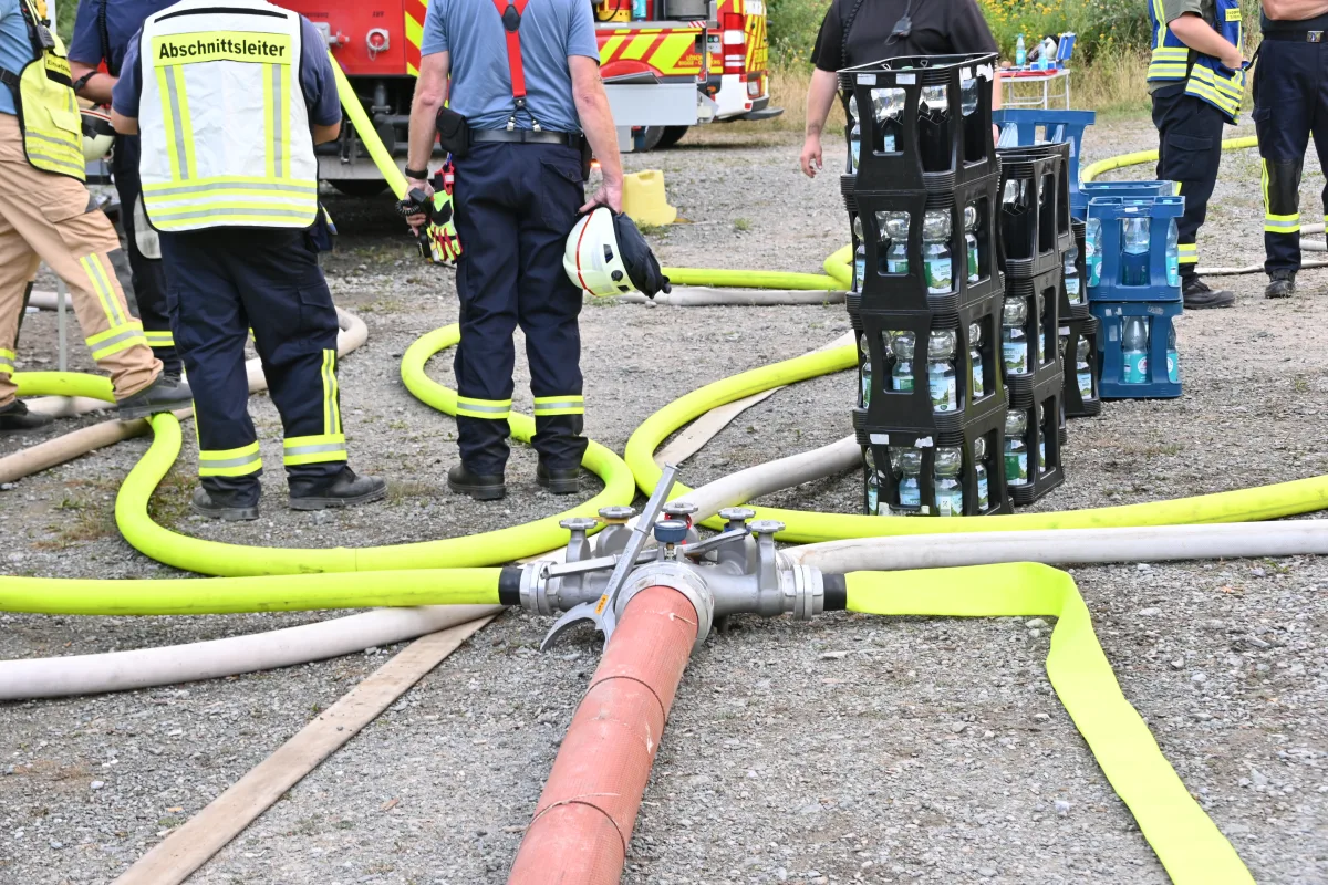Mehrere Feuerwehrleute stehen um einen Schlauchverteiler, der mit bunten Schläuchen verbunden ist. Neben ihnen stehen Kisten mit Wasserflaschen.