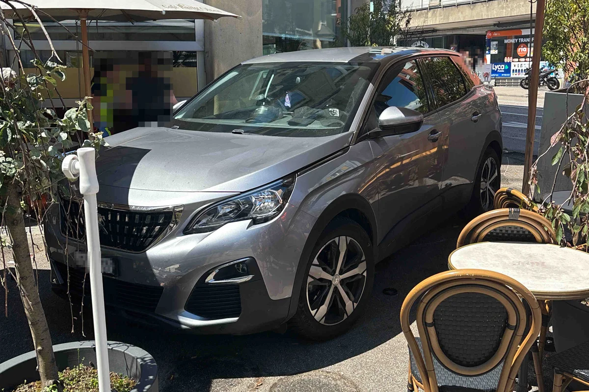Ein silbernes Auto ist in einem Straßencafé geparkt, umgeben von mehreren Tischen und Stühlen. Zwei Personen im Hintergrund sind verpixelt.