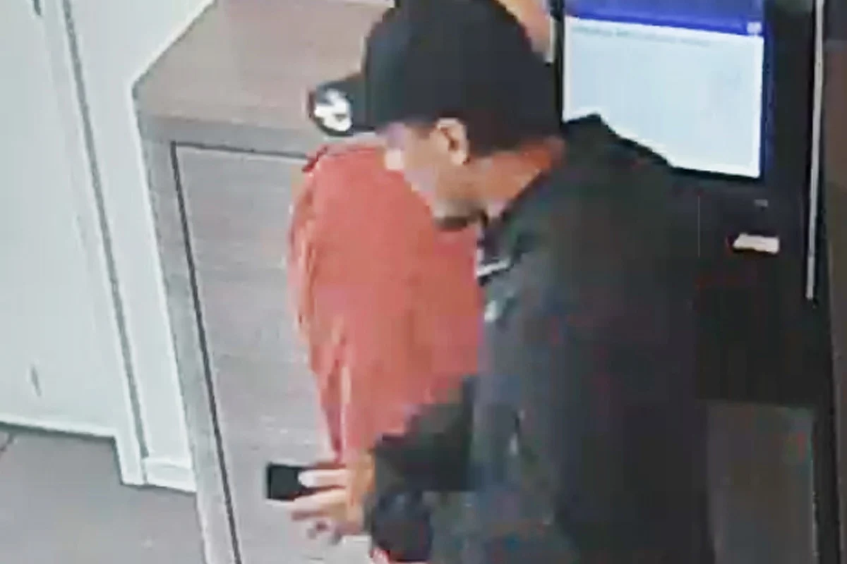 Zwei Personen stehen an einem Geldautomaten. Eine Person trägt eine rote Jacke, die andere eine schwarze Kappe und Jacke.