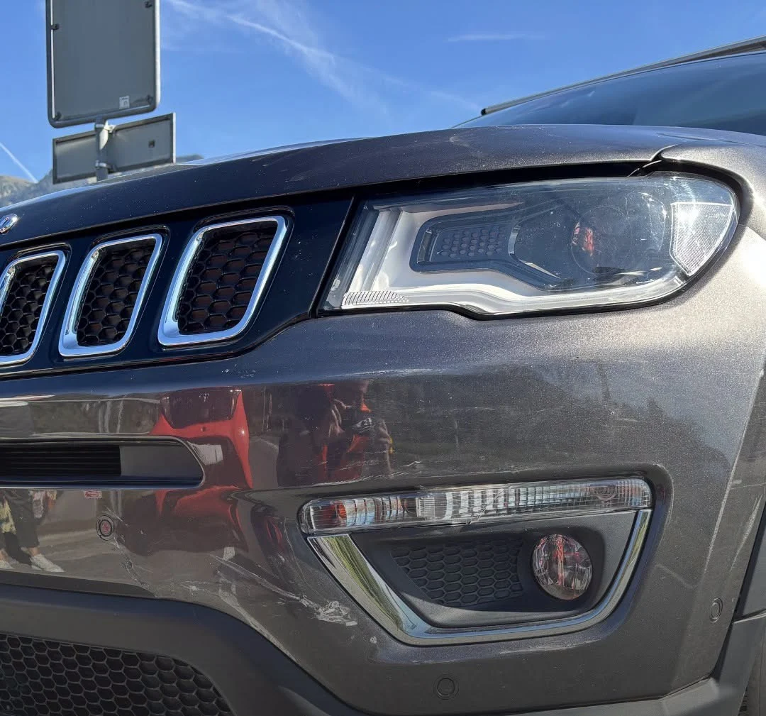 Die Nahaufnahme zeigt den Scheinwerfer eines dunklen SUV mit Kratzern auf der Stoßstange. Der Himmel ist klar blau.