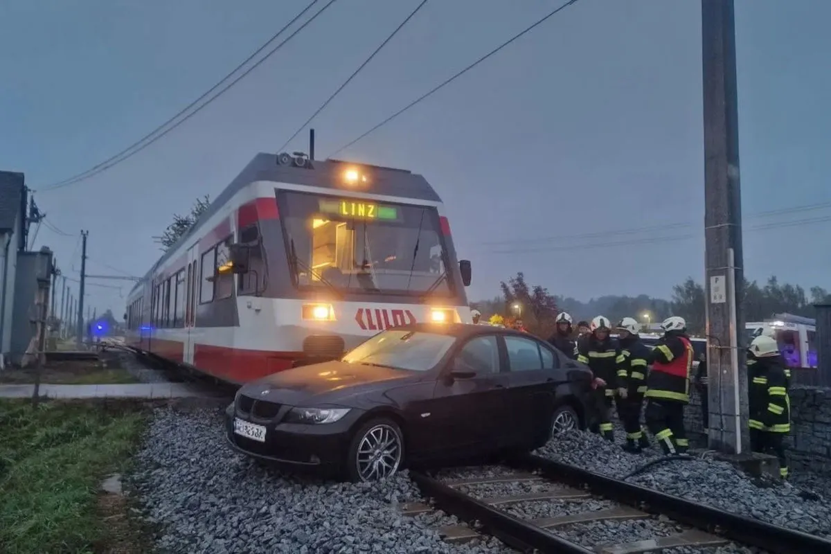 Ein Zug mit der Aufschrift 'LINZ' steht auf Gleisen, wo ein BMW hängen geblieben ist. Mehrere Feuerwehrleute sind anwesend.