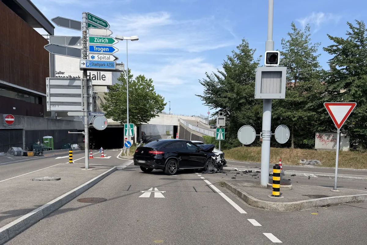 Ein schwarzes Auto mit schweren Frontschäden steht an einer Kreuzung in einer städtischen Umgebung. Straßenschilder und ein Unterführungseingang sind sichtbar.