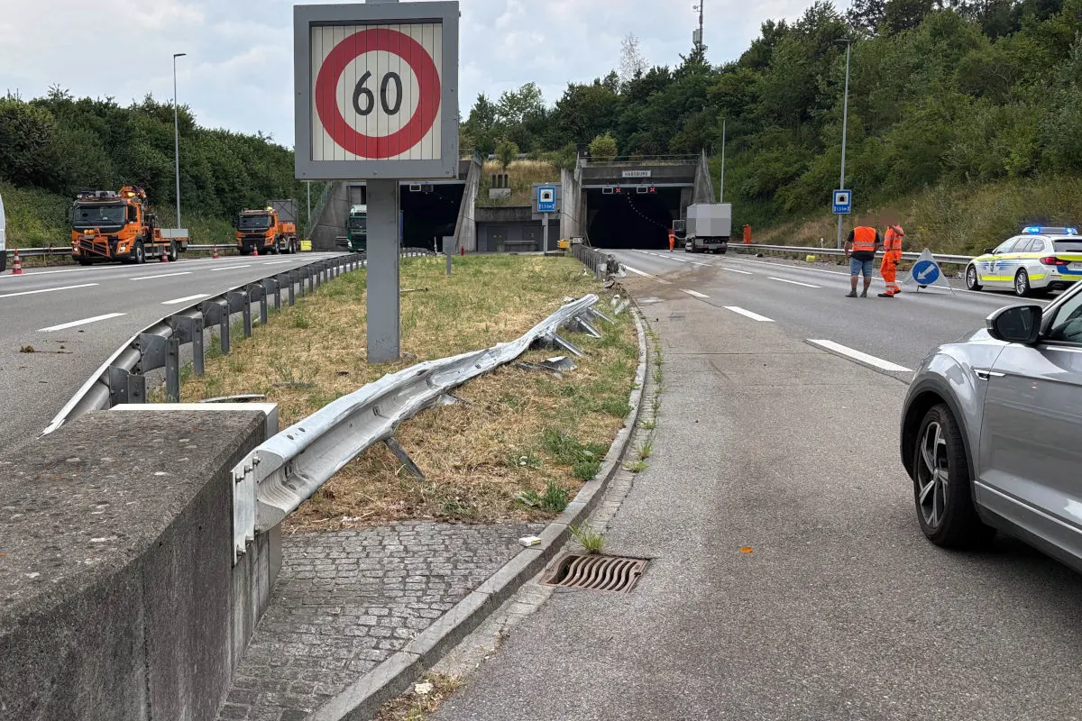 Eine Tunnelzufahrt ist zu sehen, daneben eine beschädigte Leitplanke. Ein Polizeiauto steht am Straßenrand, Arbeiter in orange warnen vor der Gefahrenstelle.
