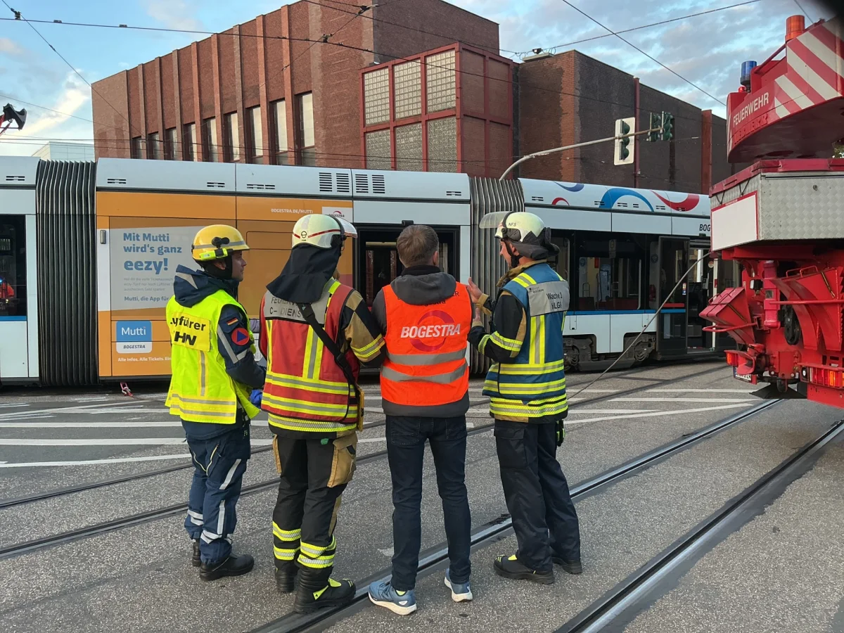 Drei Feuerwehrleute in Einsatzkleidung stehen neben einem Stadtbahnwagen und reden miteinander. Im Hintergrund ist ein rotes Feuerwehrfahrzeug zu sehen.