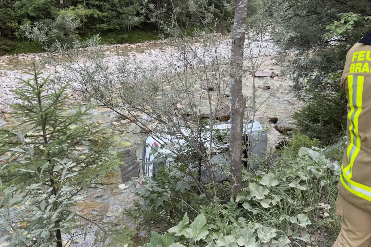 Das Bild zeigt ein Auto, das auf der Seite in einem flachen Flussbett ruht. Einige Büsche und Bäume verdecken teilweise die Sicht auf das Fahrzeug. Am rechten Bildrand ist ein Feuerwehrmann in Schutzkleidung zu sehen.