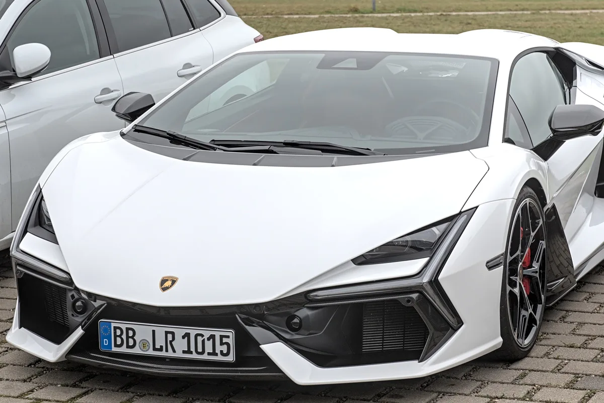 Ein weißer Sportwagen steht auf einem gepflasterten Parkplatz neben einem anderen weißen Auto. Das Kennzeichen zeigt ein deutsches Nummernschild.