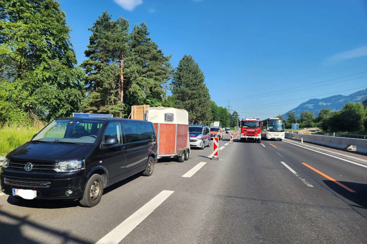 Mehrere Fahrzeuge, darunter ein schwarzer VW-Transporter mit Anhänger, ein Polizeiauto und ein Feuerwehrfahrzeug, stehen auf einer Autobahn. Berge sind im Hintergrund sichtbar.