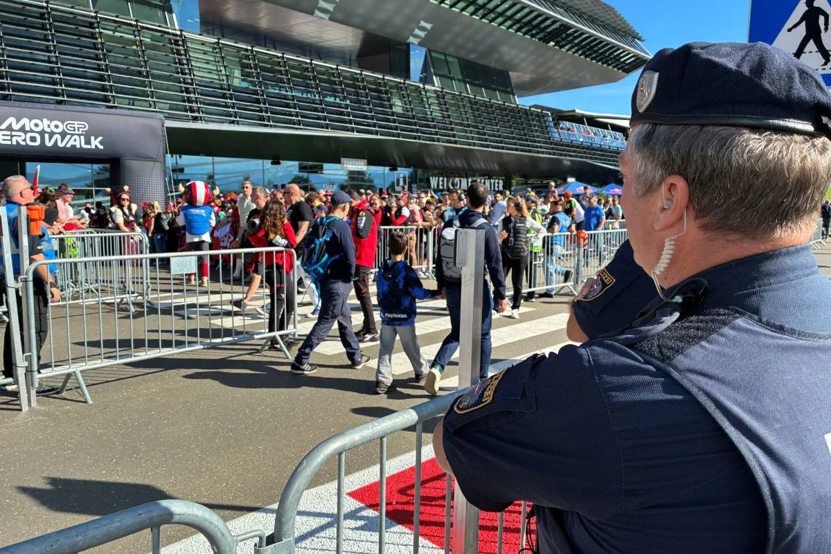 Ein Polizist mit Uniform steht vor einer Absperrung und beobachtet eine Menschenmenge bei einem Event vor einem modernen Gebäude. Es ist sonnig und der Himmel ist blau.