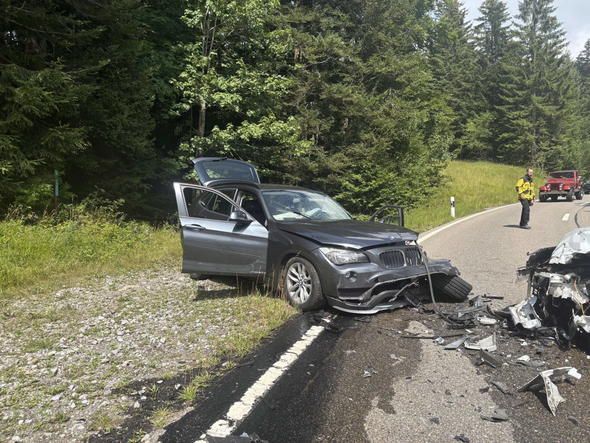 Ein beschädigter schwarzer BMW steht am Straßenrand einer Landstraße, umgeben von Bäumen. Fahrzeugtrümmer sind auf der Straße verteilt.