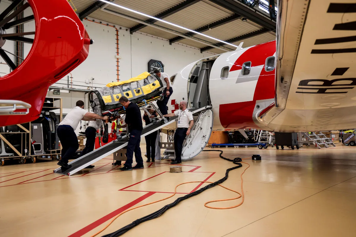 Mehrere Personen verladen eine Rettungstrage in ein rot-weißes Ambulanzflugzeug in einem Hangar.