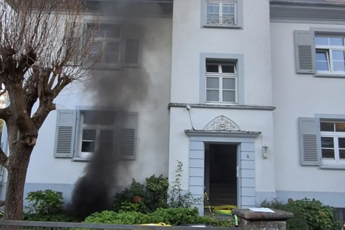 Schwarzer Rauch dringt aus dem unteren Fenster eines weißen, zweistöckigen Hauses mit grauen Fensterläden. Eine Baumreihe befindet sich links im Bild.