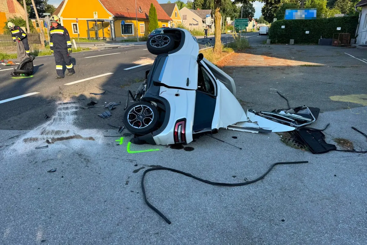 Ein weißes Auto liegt nach einem Unfall auf dem Dach, umgeben von Trümmern. Zwei Feuerwehrleute sind vor Ort zur Sicherung.