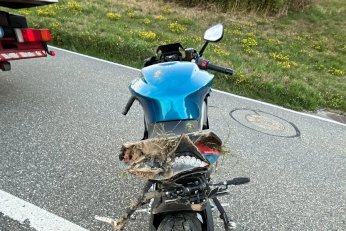 Ein blaues Motorrad mit Schäden am Heck steht auf einer Straße. Es ist nahe einem Anhänger geparkt und umgeben von Gras.