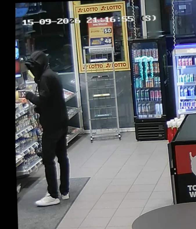 Eine vermummte Person in schwarzer Kleidung steht in einem Geschäft und greift nach Waren in einem Regal.