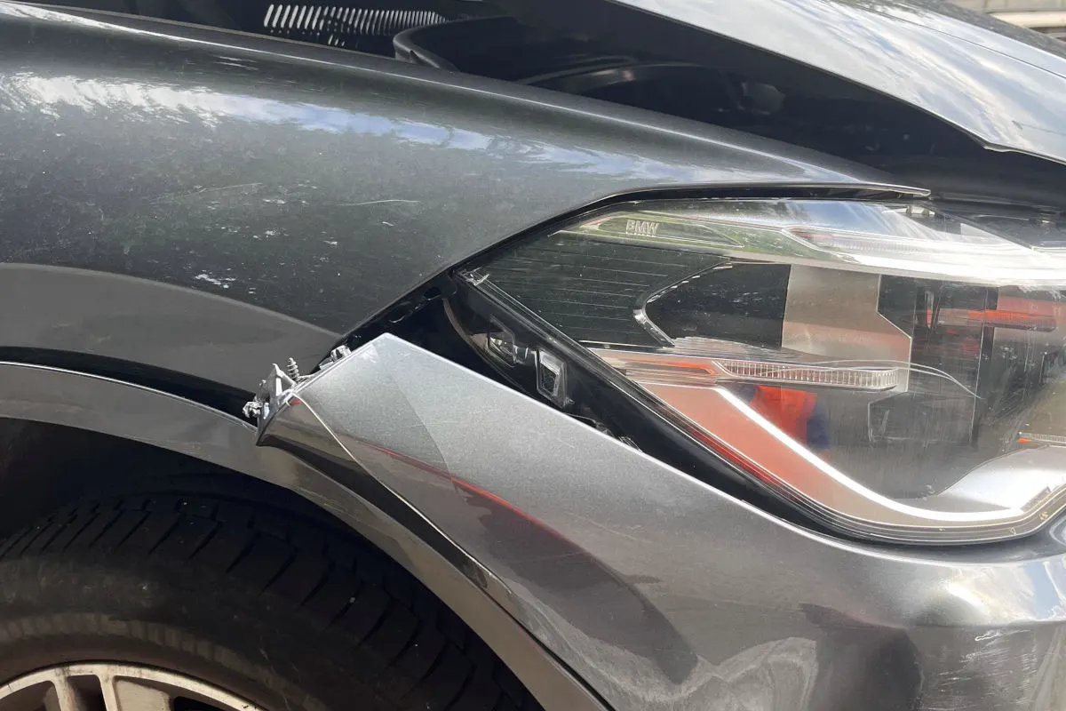 Das Bild zeigt die Nahaufnahme eines BMW mit einem beschädigten vorderen Kotflügel. Die Front weist Kratzer und Verformungen auf.