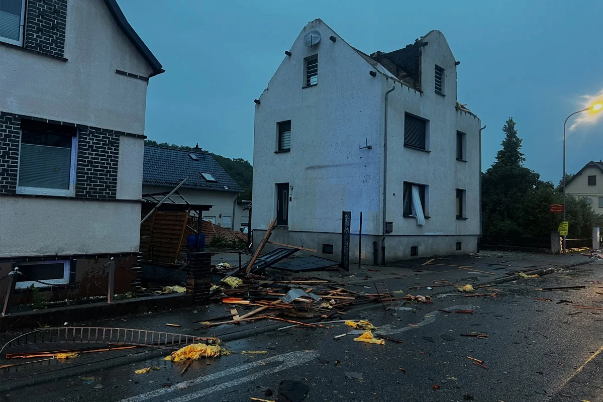 Ein Wohnhaus zeigt offensichtliche Sturmschäden, mit Trümmern und kaputten Dachziegeln auf der Straße. Der Himmel ist bedeckt und es scheint kühl zu sein.
