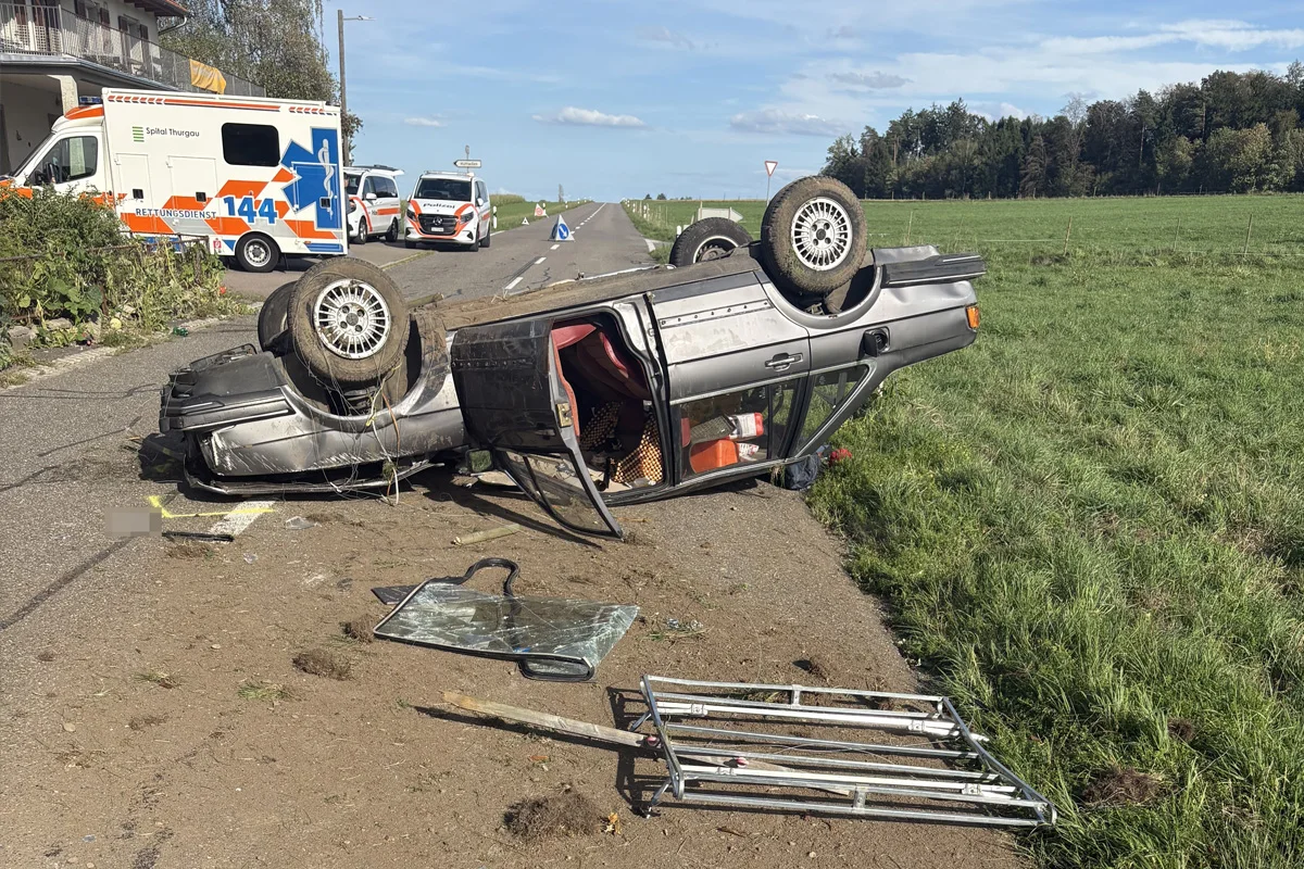 Ein Auto liegt auf dem Dach neben einer Landstraße, umgeben von Rettungsdienstfahrzeugen. Trümmerteile sind um das Fahrzeug verteilt.