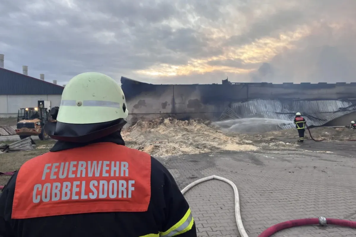 Ein Feuerwehrmann in Schutzkleidung steht vor einer brennenden Lagerhalle und beobachtet die Löscharbeiten. Im Hintergrund sind weitere Feuerwehrleute zu sehen.