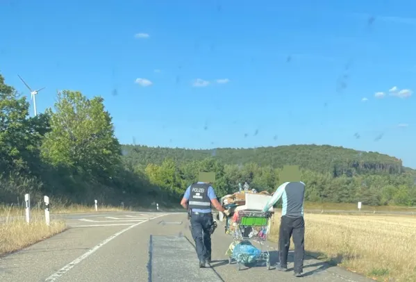 Ein Polizist geht mit einer Person und einem gepackten Einkaufswagen entlang einer Landstraße. Wälder und Felder umgeben die Szene.