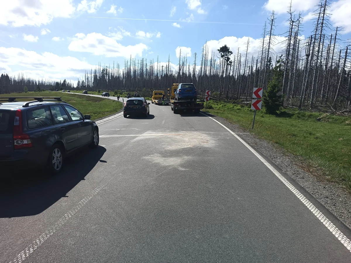 Eine ländliche Straße mit mehreren Autos, darunter ein Abschleppwagen mit einem Fahrzeug beladen. Bäume im Hintergrund.