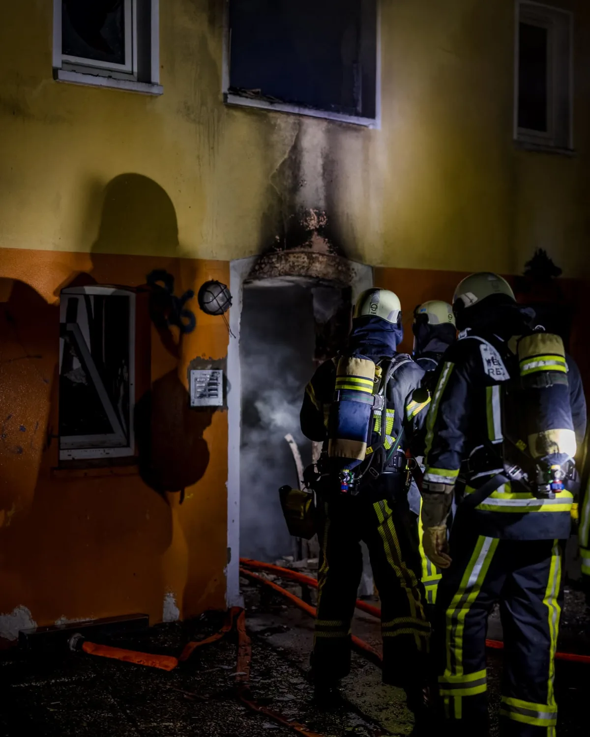 Drei Feuerwehrleute in Schutzkleidung stehen vor einem Eingang eines Hauses, das Brandspuren zeigt. Rauch ist sichtbar.