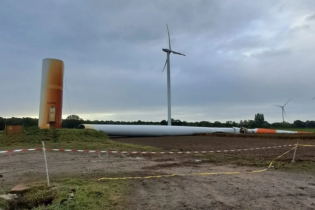 Ein Teile eines umgestürzten Windrads liegen auf einem Feld; der Himmel ist bewölkt und Absperrband grenzt den Bereich ab.