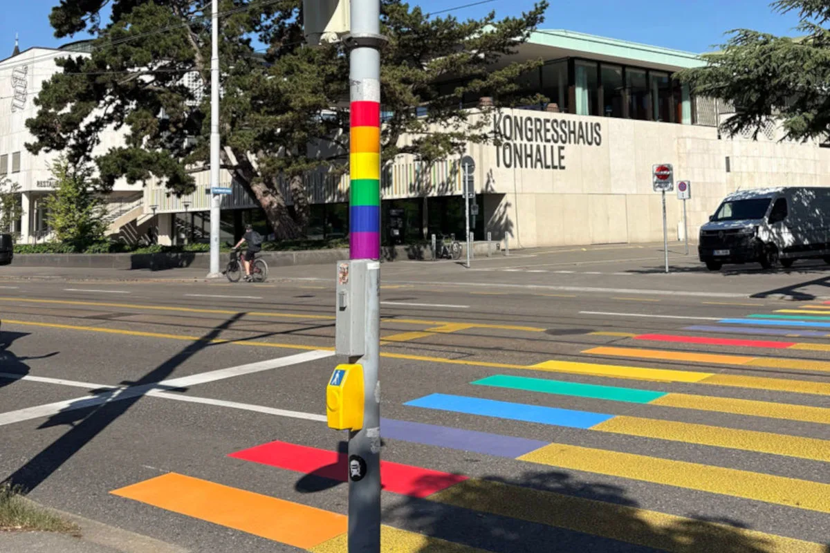 Ein farbenfroher Regenbogen-Zebrastreifen erstreckt sich über die Straße vor dem modernen Gebäude des Kongresshaus Tonhalle an einem sonnigen Tag.