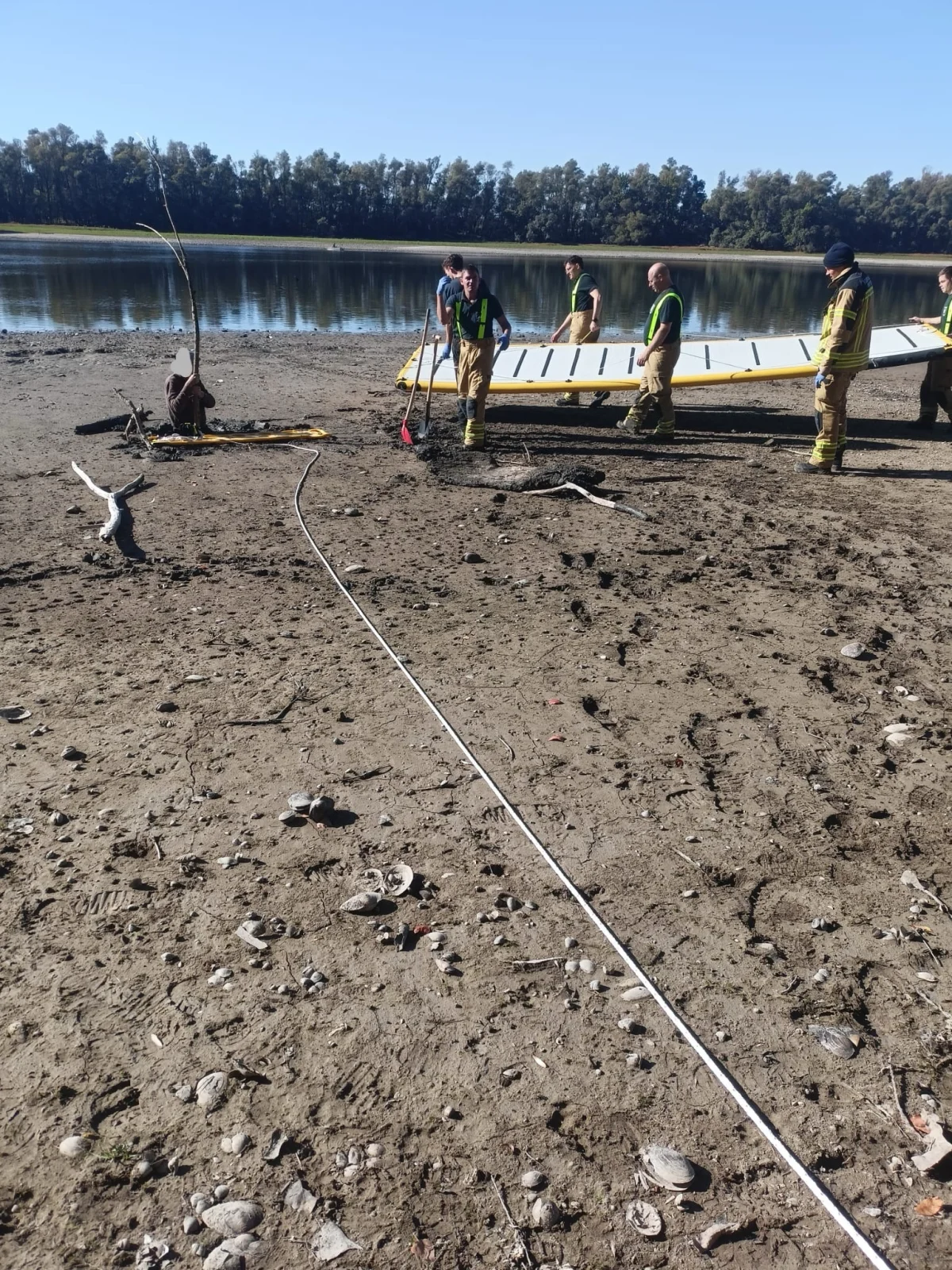 Mehrere Einsatzkräfte stehen auf einem trockenen Flussufer und bergen mit Werkzeugen ein zersplittertes Boot. Der Hintergrund zeigt Wasser und Bäume.