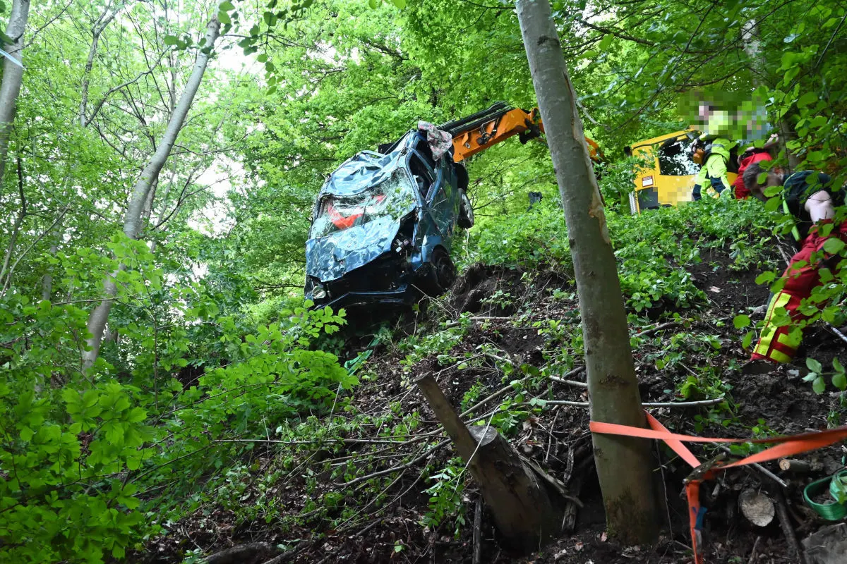 Ein stark beschädigtes Auto hängt an einem bewaldeten Hang, während Rettungspersonal sichtbar ist. Ein Bagger unterstützt bei der Bergung.