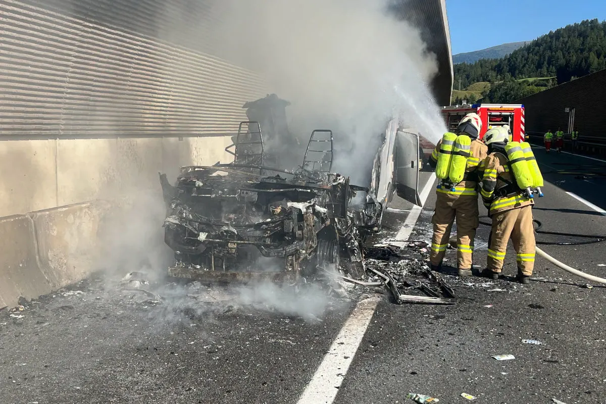 Ein stark beschädigtes Fahrzeug am Fahrbahnrand der Autobahn wird von zwei Feuerwehrleuten gelöscht. Dicker Rauch steigt auf, während Trümmer auf der Straße liegen.