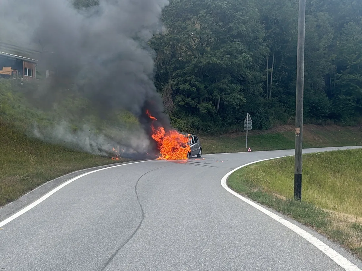 Ein Auto steht in Flammen auf einer kurvigen Straße, dichter Rauch steigt auf und eine Warnbarke steht neben der Fahrbahn.