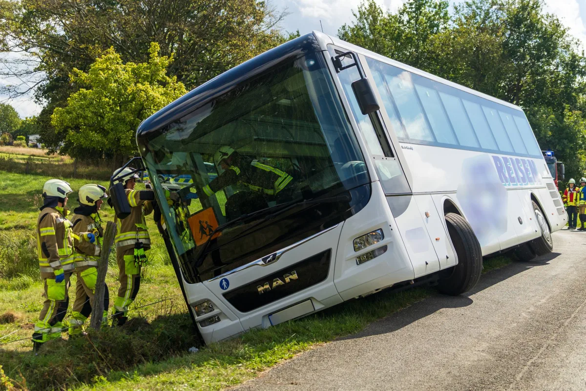 Ein weißer Reisebus steckt schräg in einem Graben an einer Landstraße, während mehrere Feuerwehrleute in gelben Schutzanzügen am Bus arbeiten. Im Hintergrund sind Bäume und eine Wiese zu sehen.