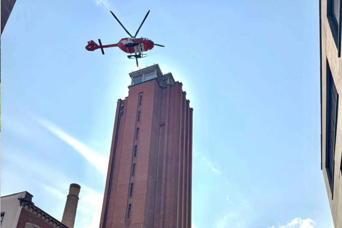 Ein roter Rettungshubschrauber fliegt am blauen Himmel über einen hohen Backsteinturm in einer städtischen Umgebung.