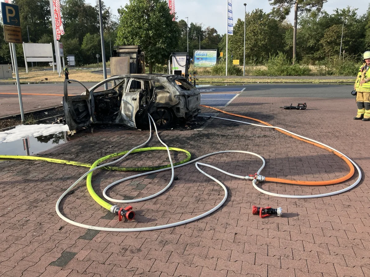 Ein ausgebranntes Auto steht auf einem Parkplatz, umgeben von Feuerwehrschläuchen. Ein Feuerwehrmann ist in der Nähe sichtbar.