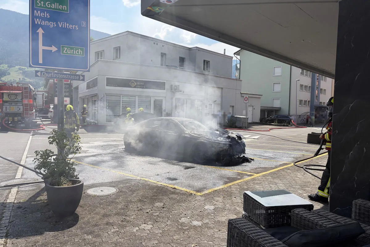 Ein ausgebranntes Auto wird auf einem Parkplatz von Feuerwehrleuten gelöscht. Im Hintergrund sind ein Gebäude und ein Feuerwehrauto zu sehen.