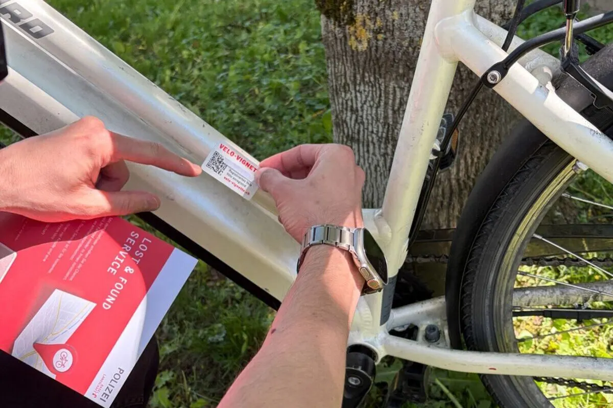 Ein Mann mit Armbanduhr befestigt im Freien eine Velo-Vignette an einem weißen Fahrradrahmen. Dabei hält er ein Dokument mit dem Titel 'Lost & Service Found'.
