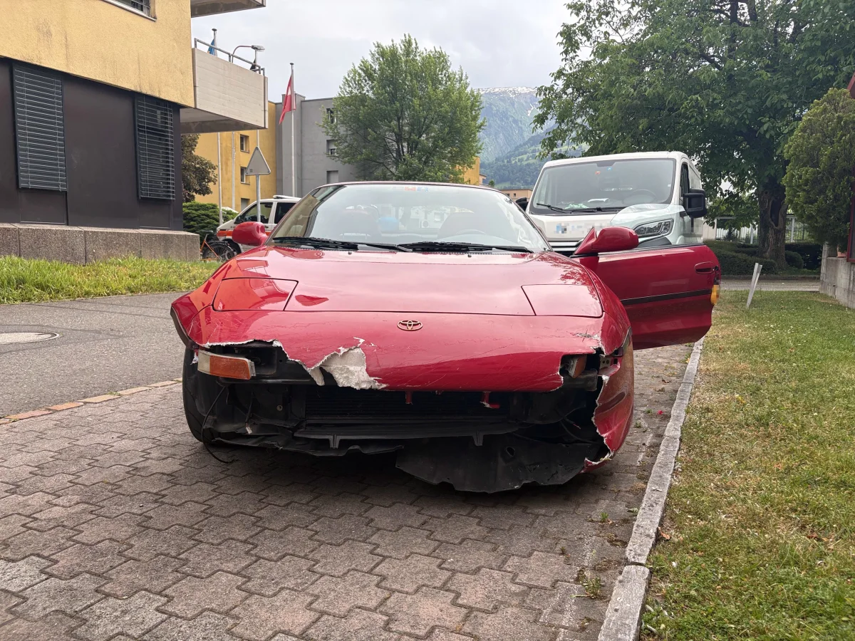 Ein roter Toyota mit deutlichem Frontschaden steht auf einem Gehweg. Im Hintergrund sind Wohngebäude und ein Lieferwagen sichtbar.