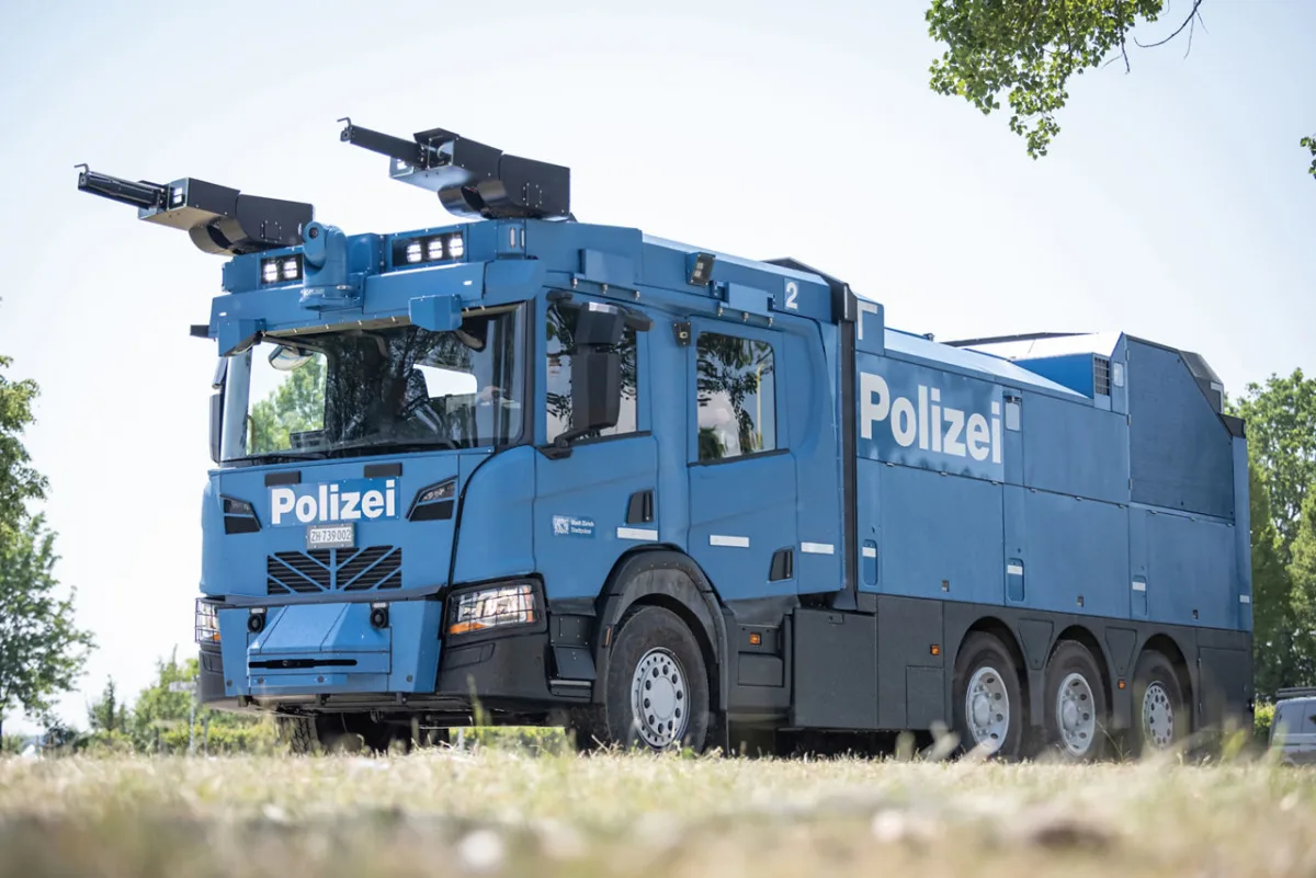Ein großer blauer Wasserwerfer der Polizei Zürich steht auf einem Grasfeld. Das Fahrzeug zeigt klare Polizei-Markierungen und ist bei Tageslicht zu sehen.