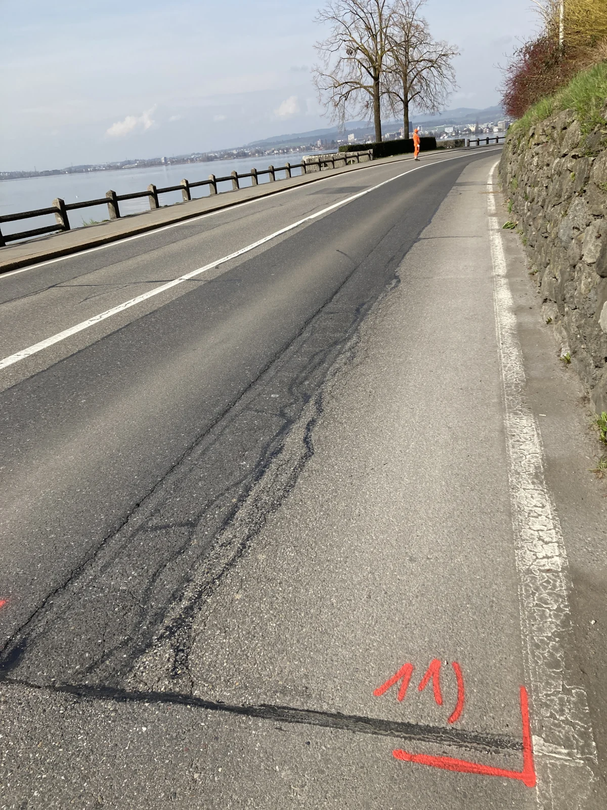 Eine leere Straße am Ufer des Bodensees zeigt rote Markierungen. Ein Arbeiter in orangefarbener Kleidung geht den Gehweg entlang.